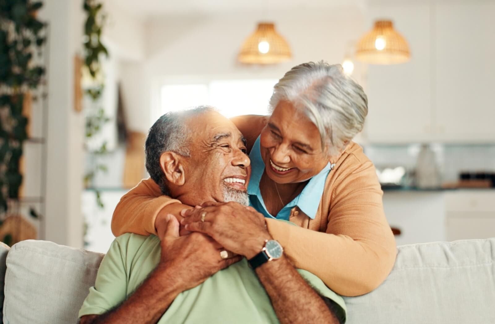 A smiling older adult leans over and embraces their spouse, who is sitting on a couch.
