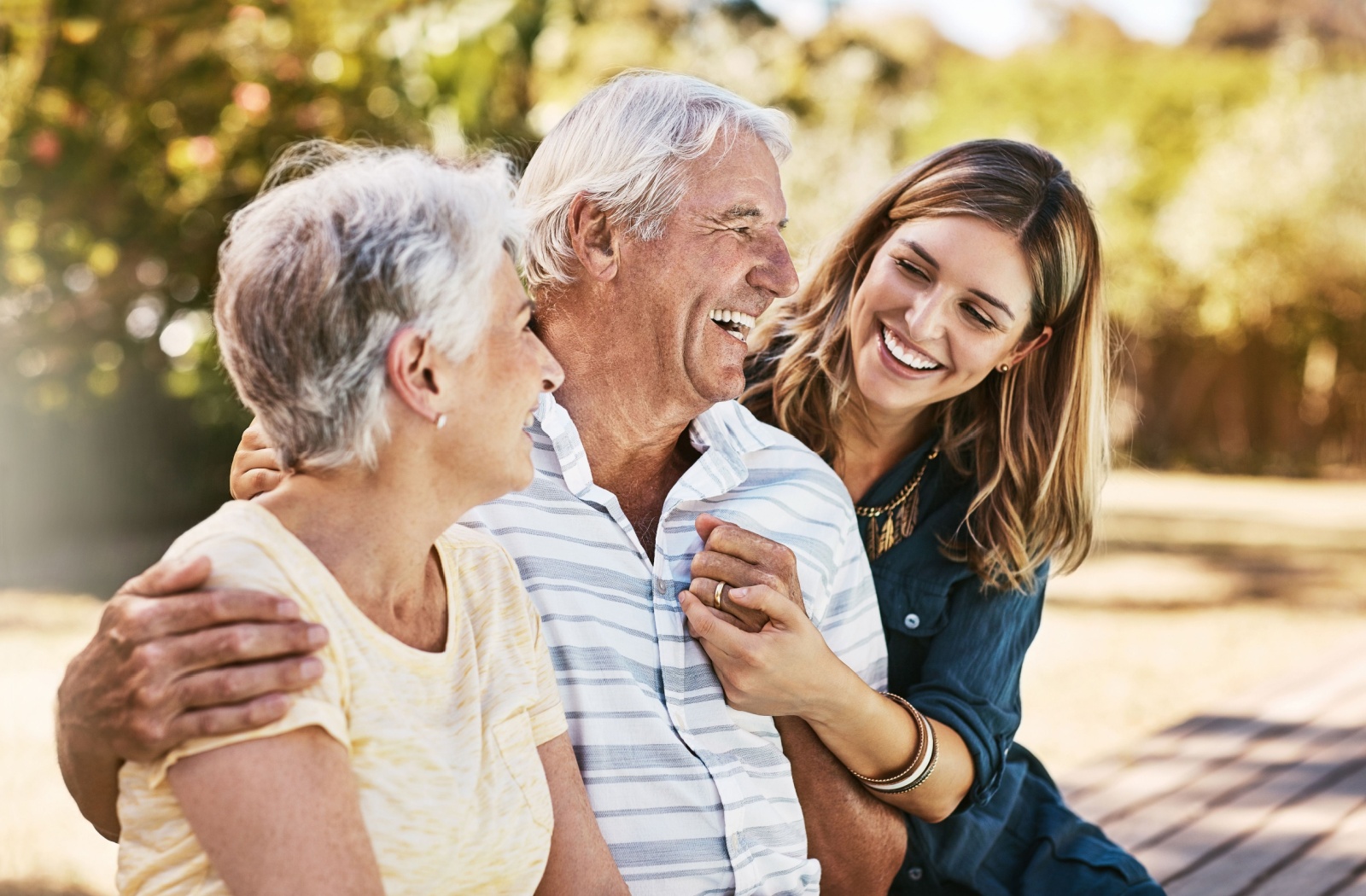 An adult child embraces their older parents while sitting on a bench outside.