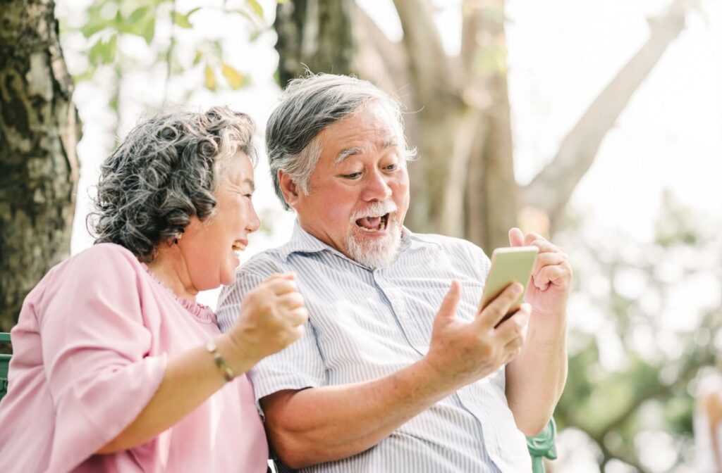 2 older adults sitting smiling and using a smartphone.