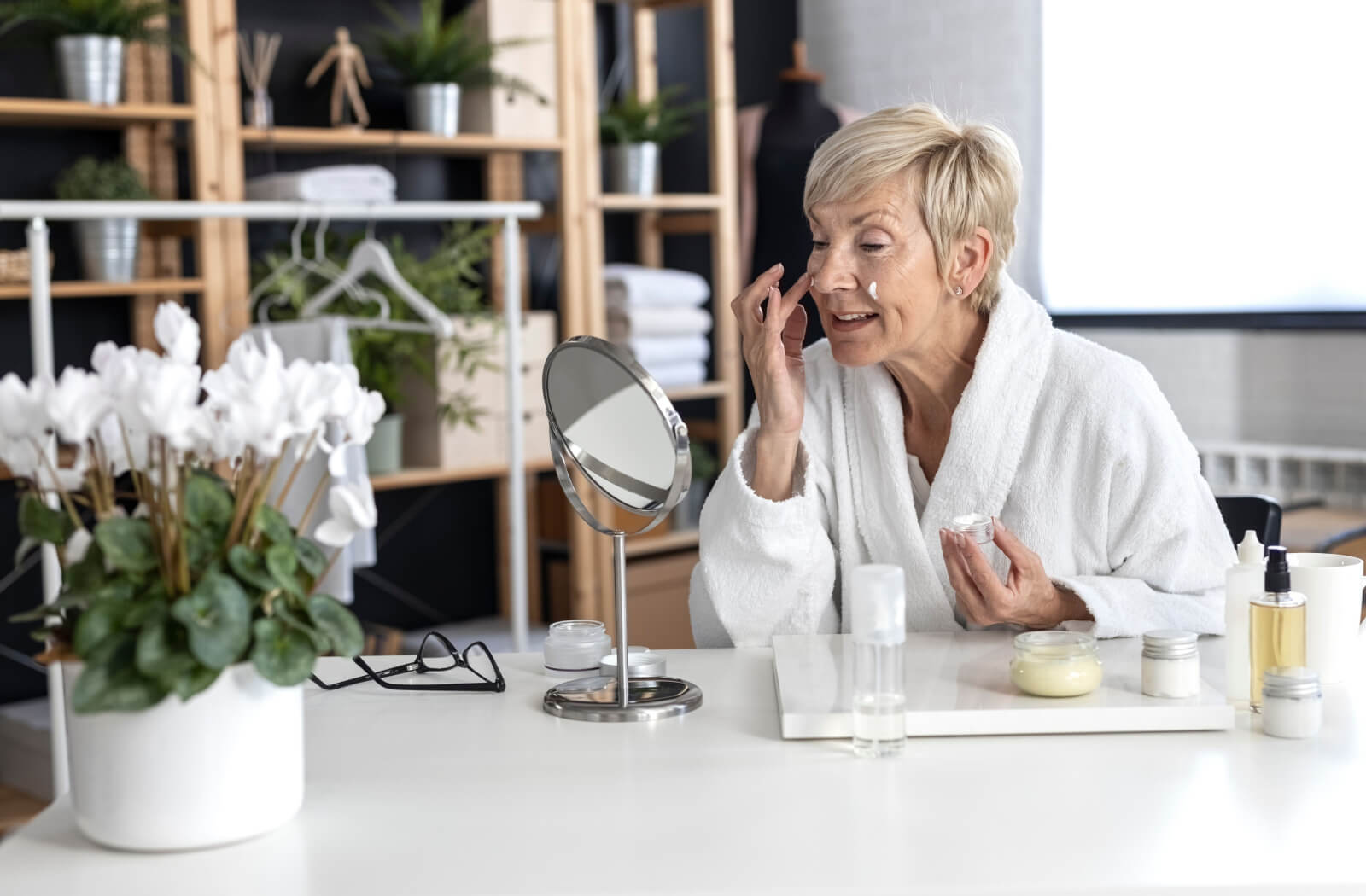 A woman diligently applies skincare products to her face, standing in front of a mirror, embracing a nurturing routine for her skin.