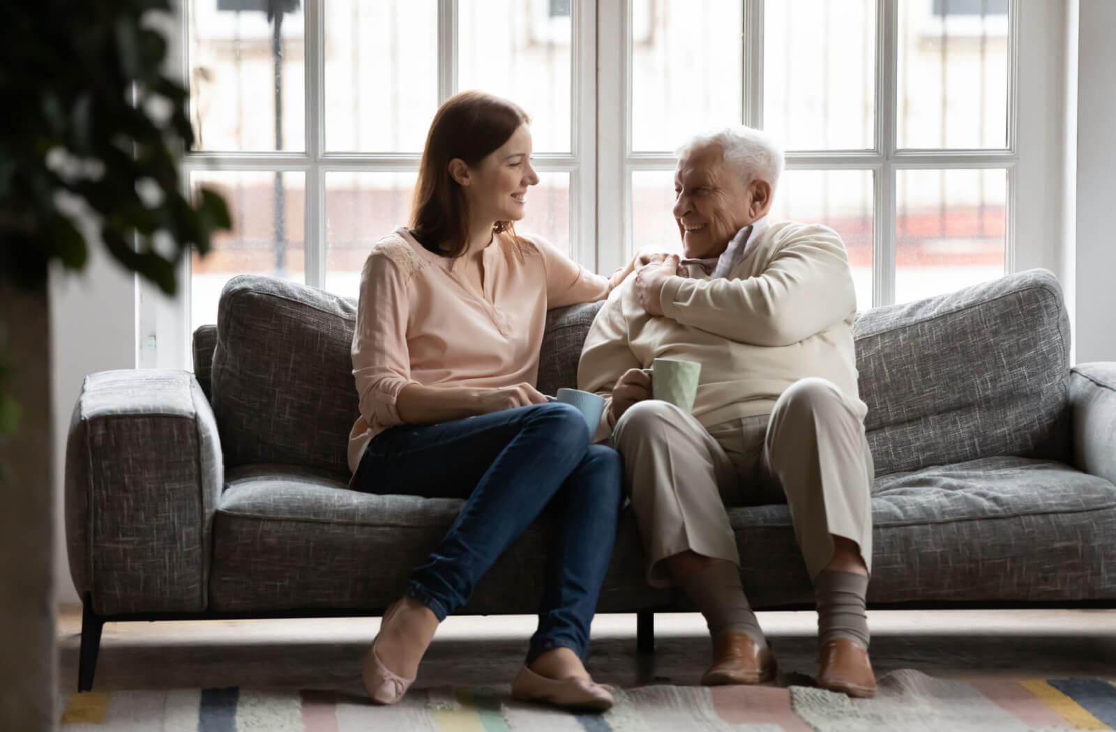 An adult daughter and her father with dementia, sit on a couch, discussing the prospect of moving to an assisted living facility over a cup of coffee, navigating a difficult but necessary conversation with care and understanding.