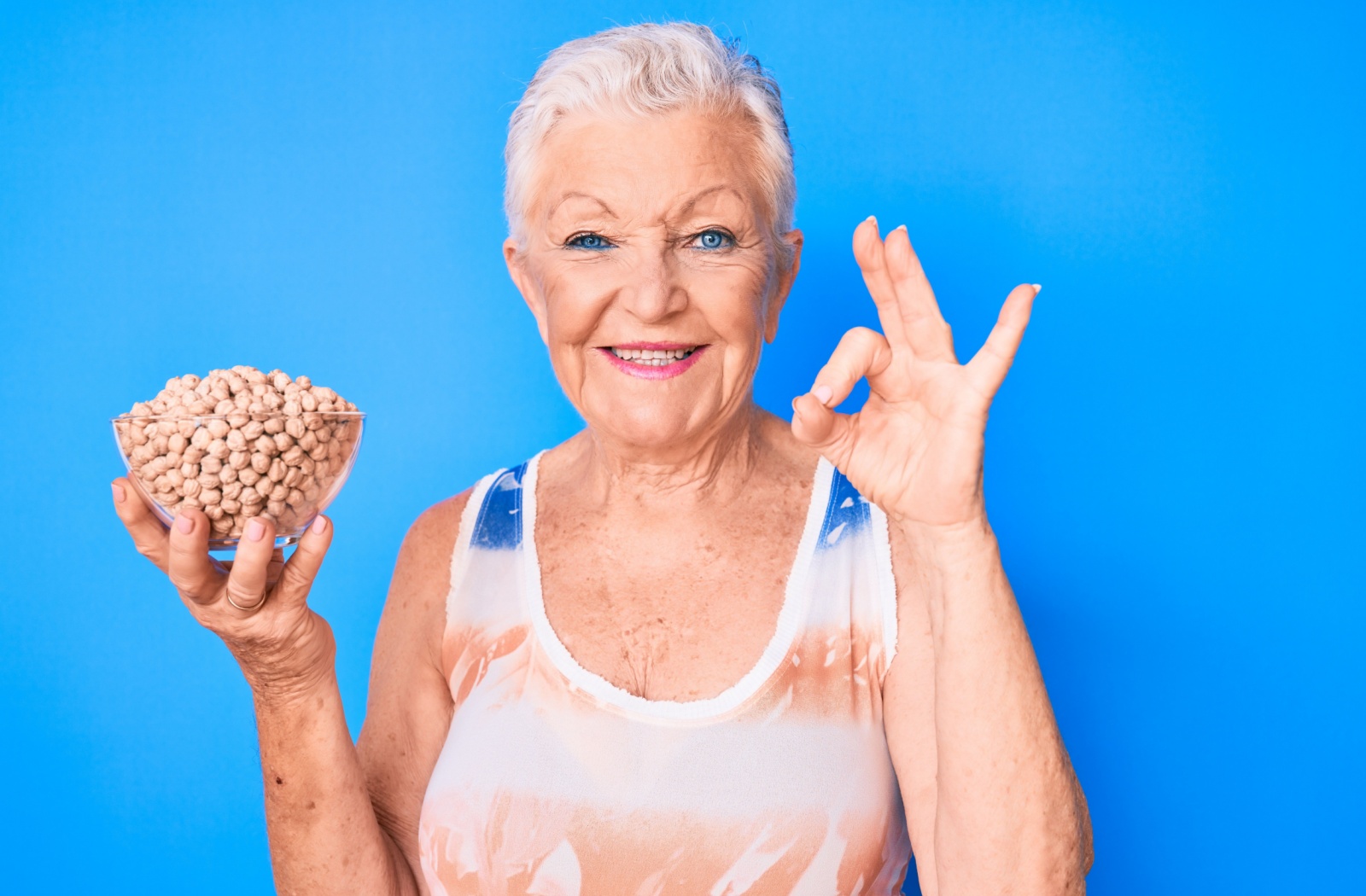 An older adult standing in front of a blue background with a bowl of dried chickpeas in their right hand and an okay hand sign with their left hand.
