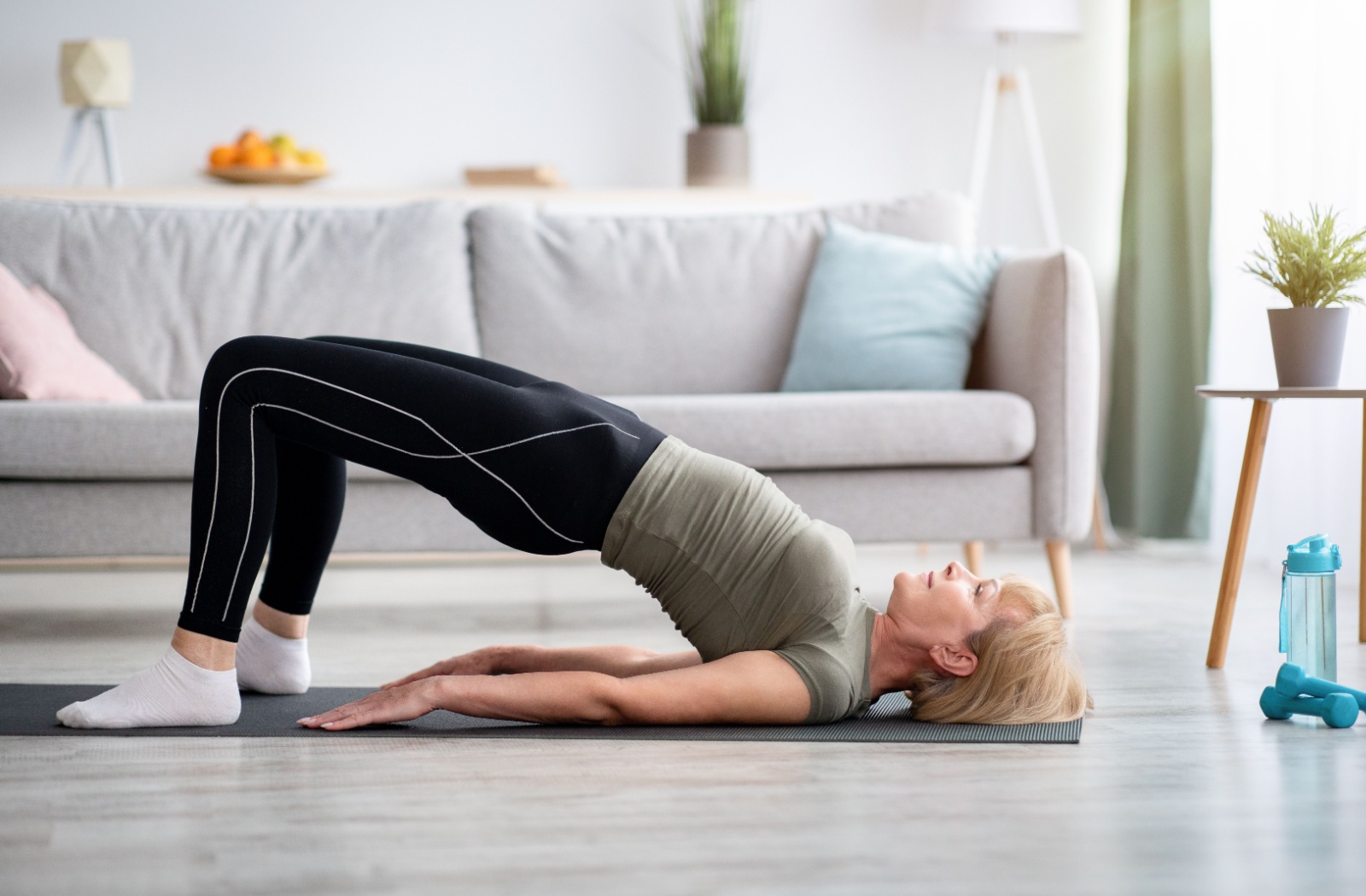 An older adult in a sunlit living room on a yoga mat, performing a bridge exercise to strengthen their core.
