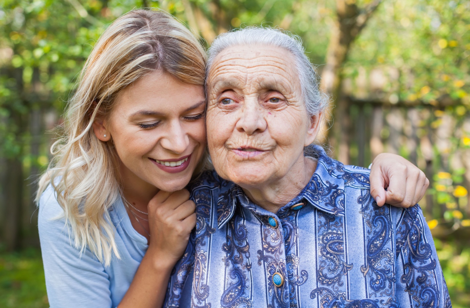 A smiling adult child embracing her mother with both hands on her shoulders while outdoors.