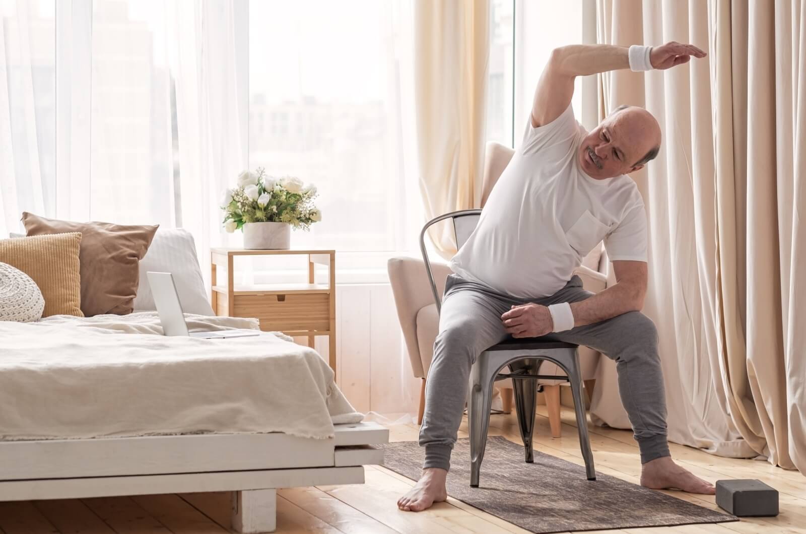 An older adult sitting on a chair while stretching their right arm overhead.