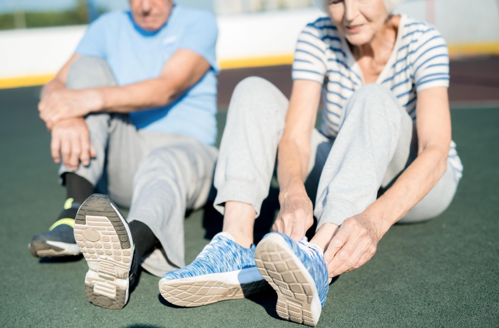 Two seniors sit on the ground while adjusting the laces on their shoes to ensure proper footwear for going on a walk
