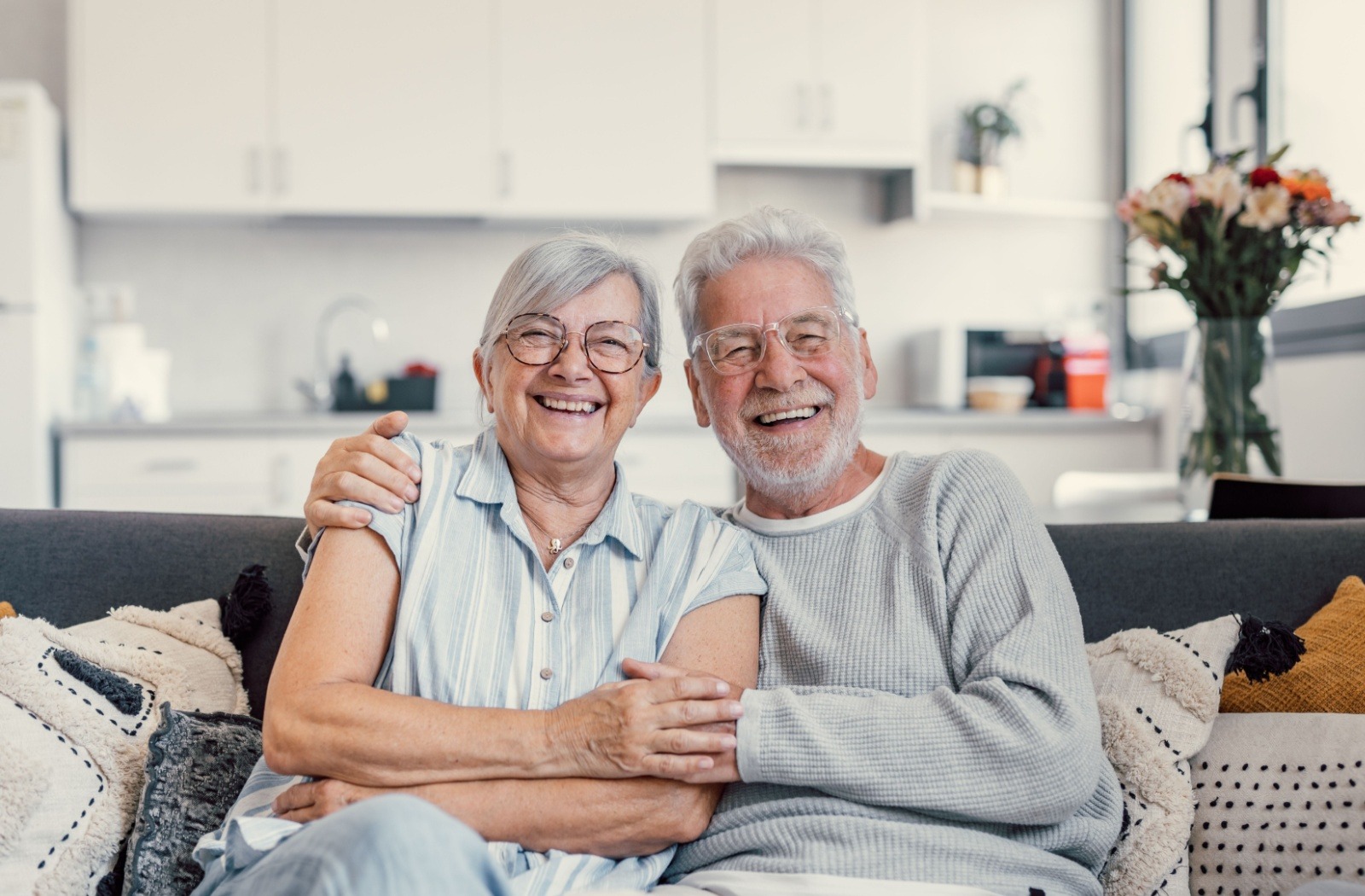 A senior couple sits on a couch together holding hands after downsizing into a senior living community.
